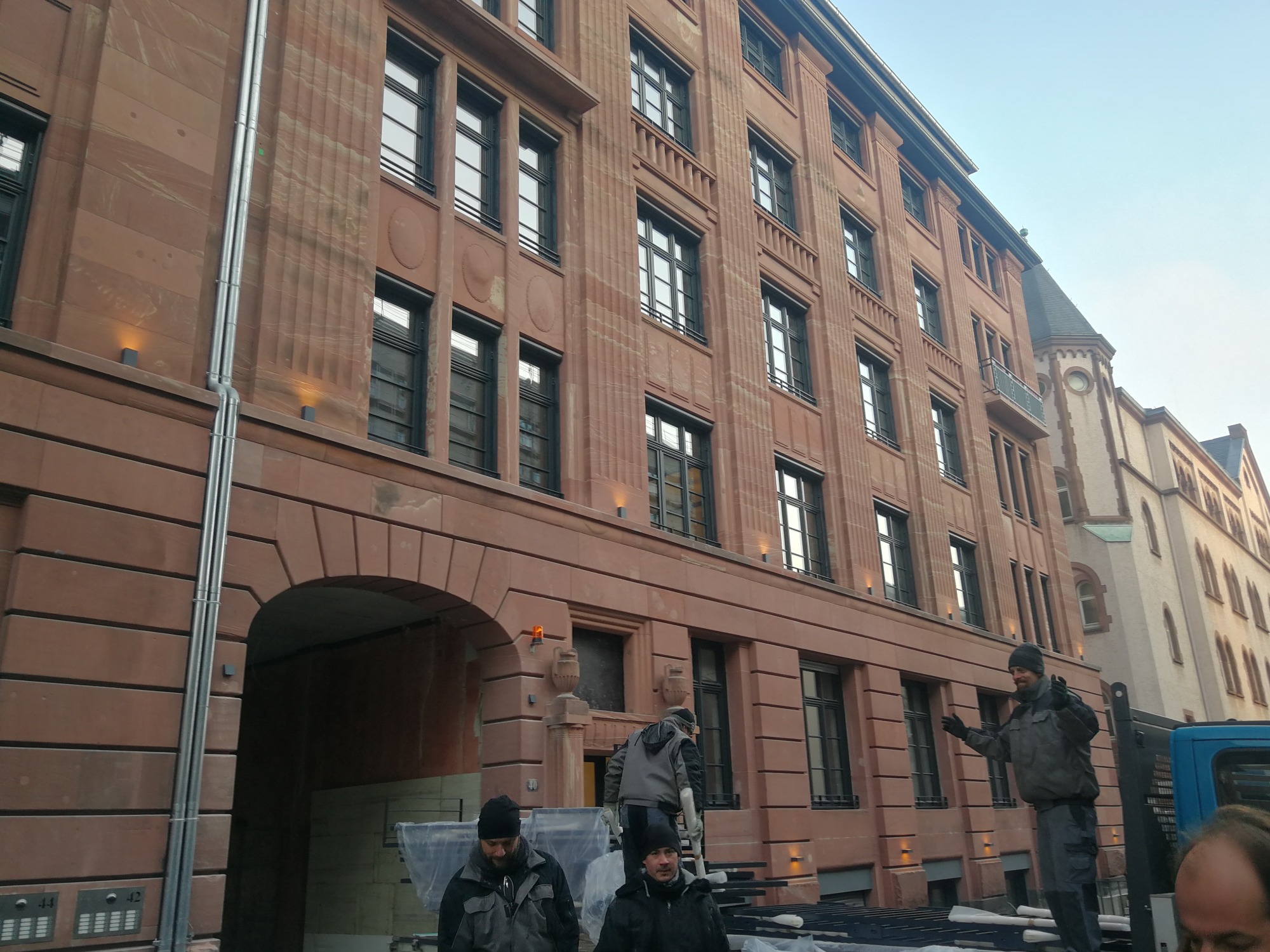 Pracownicy na rusztowaniu montują oświetlenie na elewacji budynku z czerwonej cegły, widoczne rury spustowe i lampy elewacyjne.