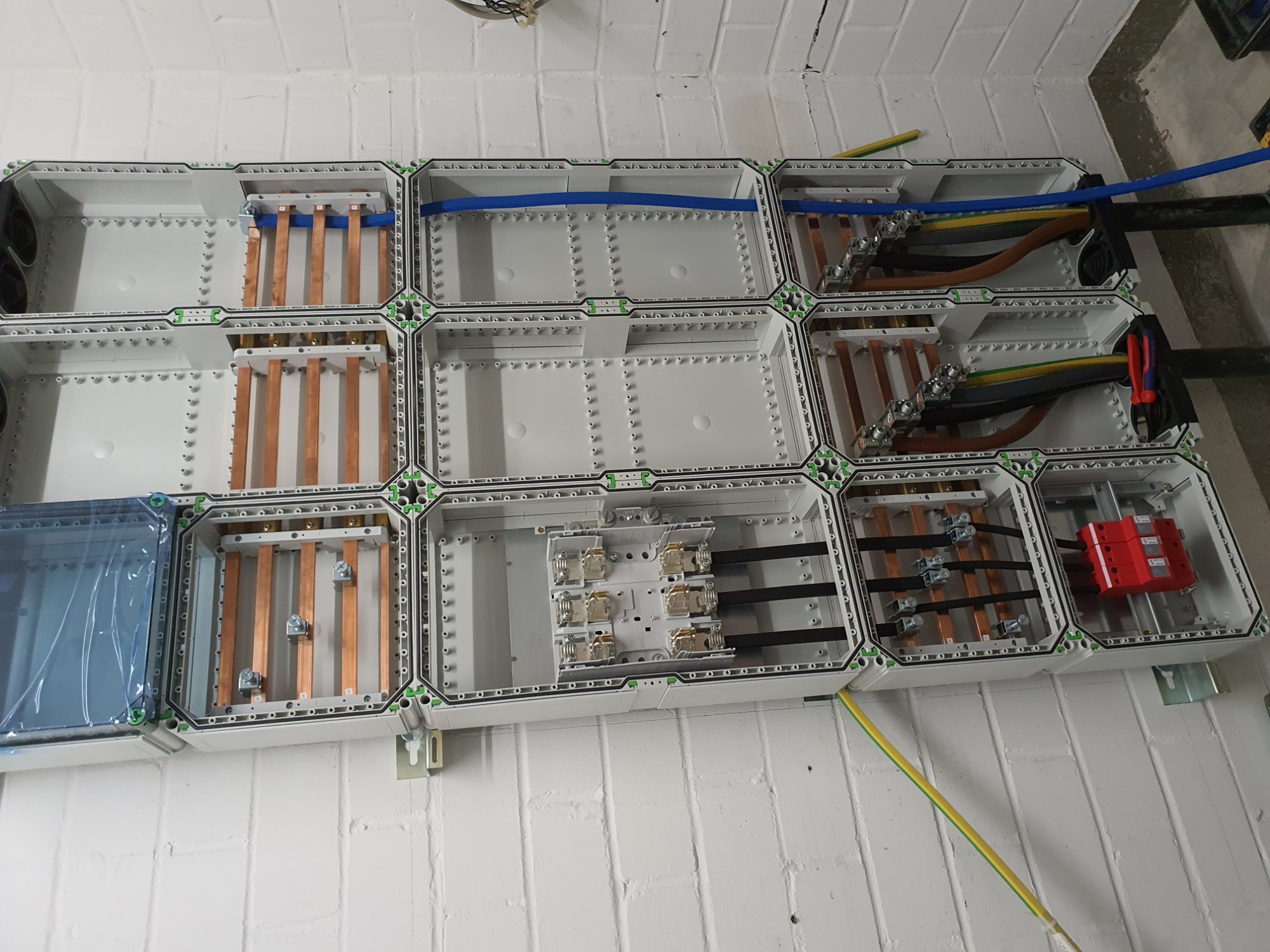 Rozdzielnica elektryczna modułowa, widoczne szyny zbiorcze miedziane, bezpieczniki, przewody w kolorach: czarnym, brązowym, żółto-zielonym i niebieskim, zamontowana na białej ścianie z cegły.