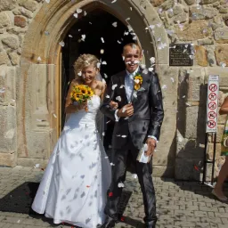 Uśmiechnięta para młoda wychodzi z kamiennego kościoła, obsypywana białymi płatkami kwiatów; panna młoda trzyma bukiet słoneczników, a pan młody ma na twarzy kilka płatków.