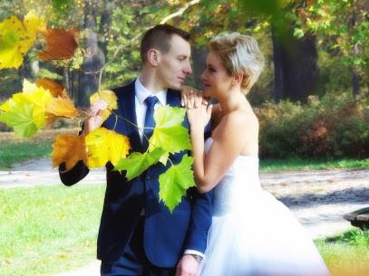 Para młoda w jesiennej scenerii parku; panna młoda w białej sukni, pan młody w granatowym garniturze, oboje patrzą na siebie trzymając gałązkę z kolorowymi liśćmi.