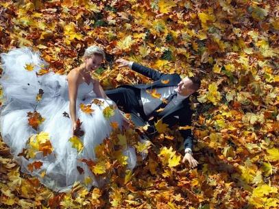 Para młoda leżąca wśród jesiennych liści, panna młoda w białej sukni z tiulu, pan młody w czarnym garniturze, uśmiechnięci, liście klonowe w odcieniach żółci i pomarańczy.