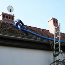 Pracownik w niebieskim kombinezonie rozprowadza izolację piankową za pomocą niebieskiego węża na dachu domu z ceglanym kominem i anteną satelitarną, drabina oparta o rynnę.