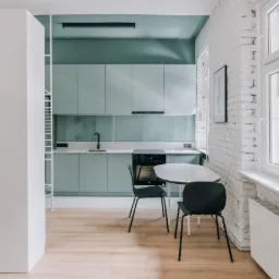 Jasna kuchnia z miętową zabudową, białymi ścianami z cegły i drewnianą podłogą, uzupełniona czarnymi krzesłami i okrągłym stołem.