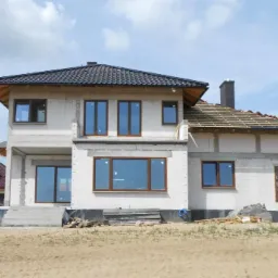 Dwukondygnacyjny budynek w trakcie budowy, z częściowo ukończonym dachem krytym czarną dachówką, widoczne przygotowania do dalszych prac dekarskich na drugiej połowie dachu, okna w brązowych ramach...