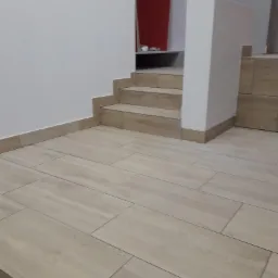 Ułożona podłoga z płytek imitujących drewno, prowadząca do schodów z wykończonymi krawędziami i białym filarem, widok na czerwony element dekoracyjny w tle.