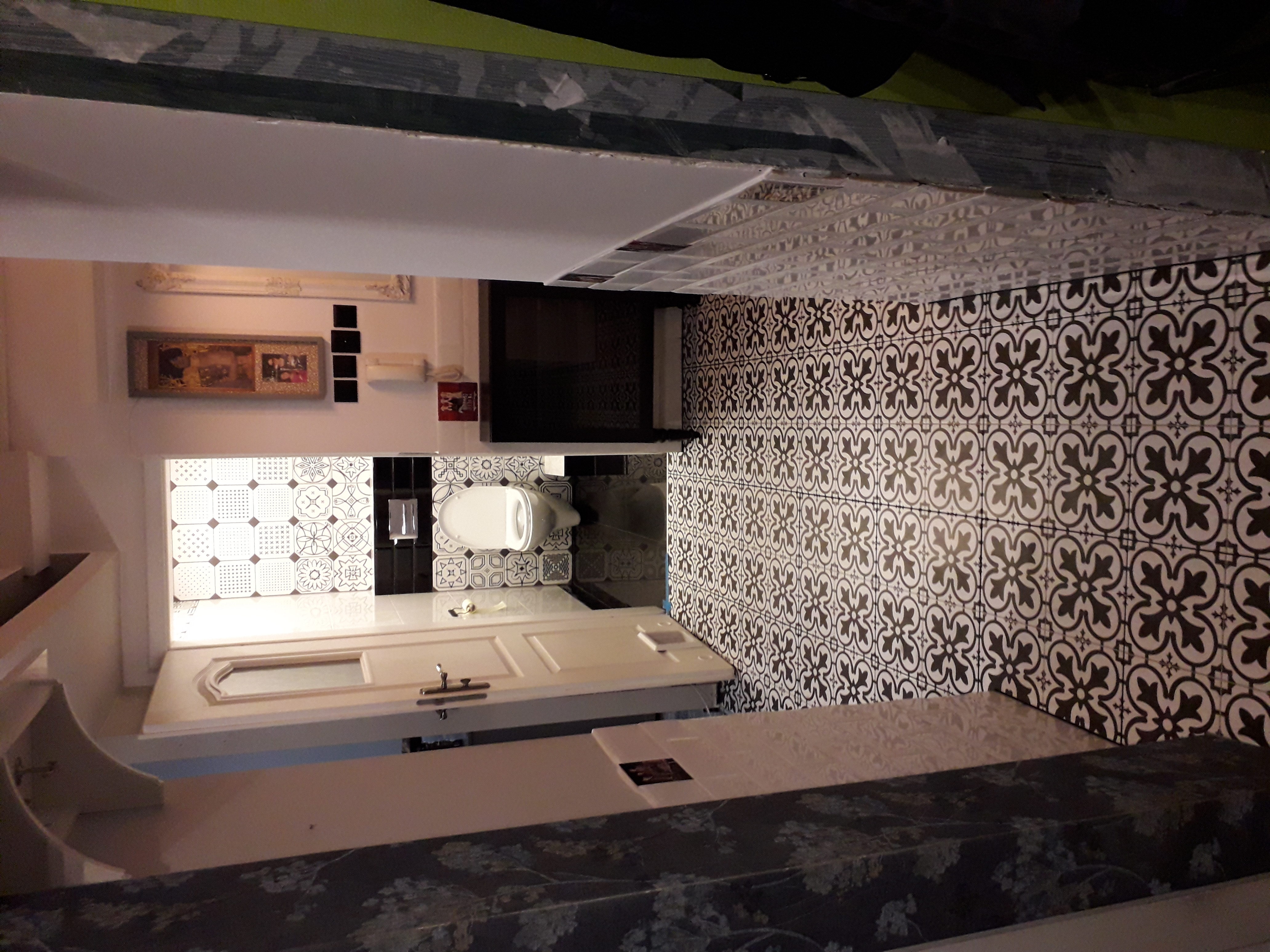 Wykończona łazienka z białą toaletą, wzorzystymi płytkami na podłodze i ścianach, oraz białą zabudową pod umywalką, widok z góry z perspektywy drzwi.