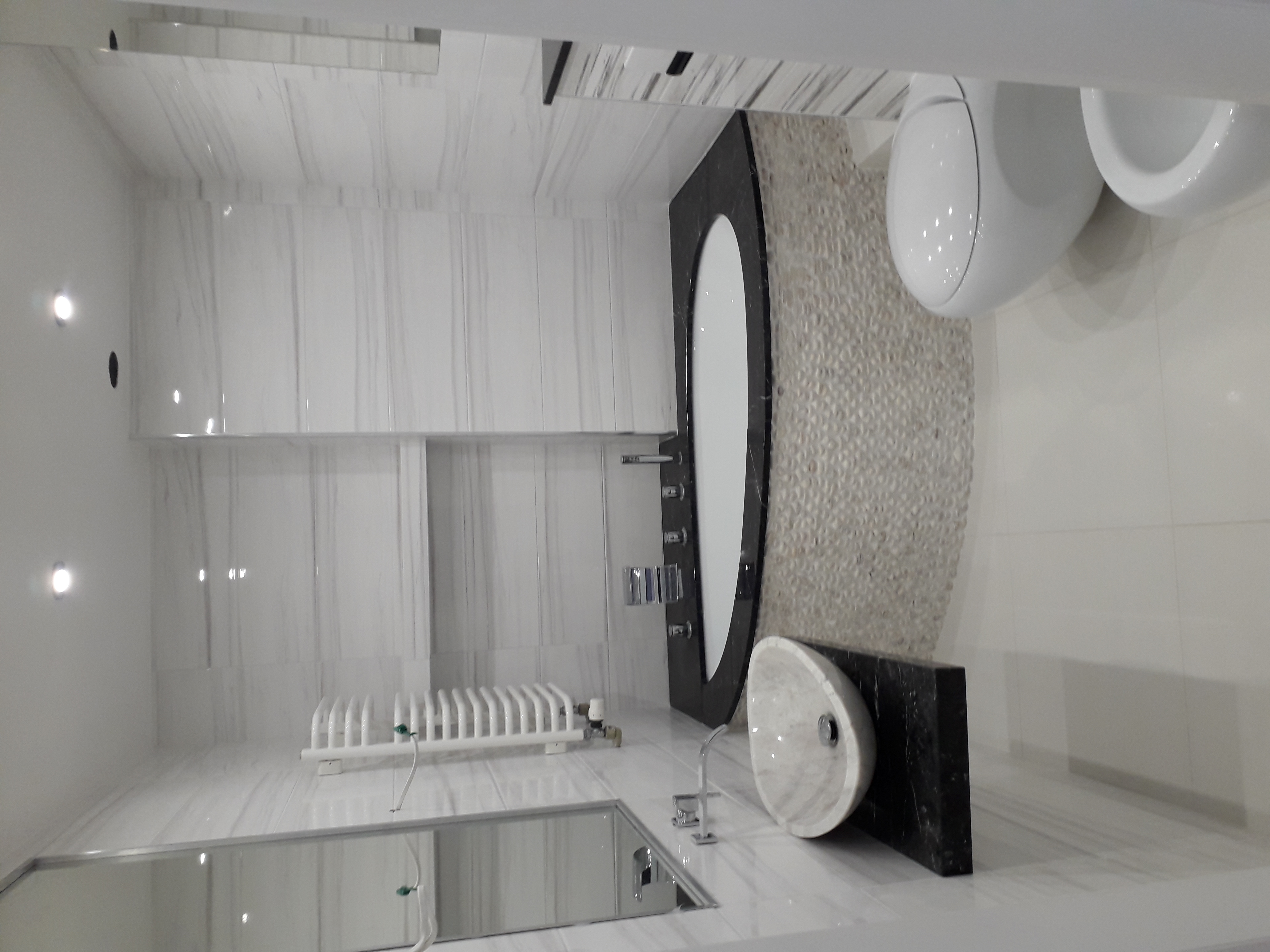 Elegancka łazienka w bieli i czerni z marmurowymi płytkami, owalnym lustrem z czarną ramą, umywalką nablatową z marmuru, i ścianą pokrytą mozaiką z drobnych kamieni.