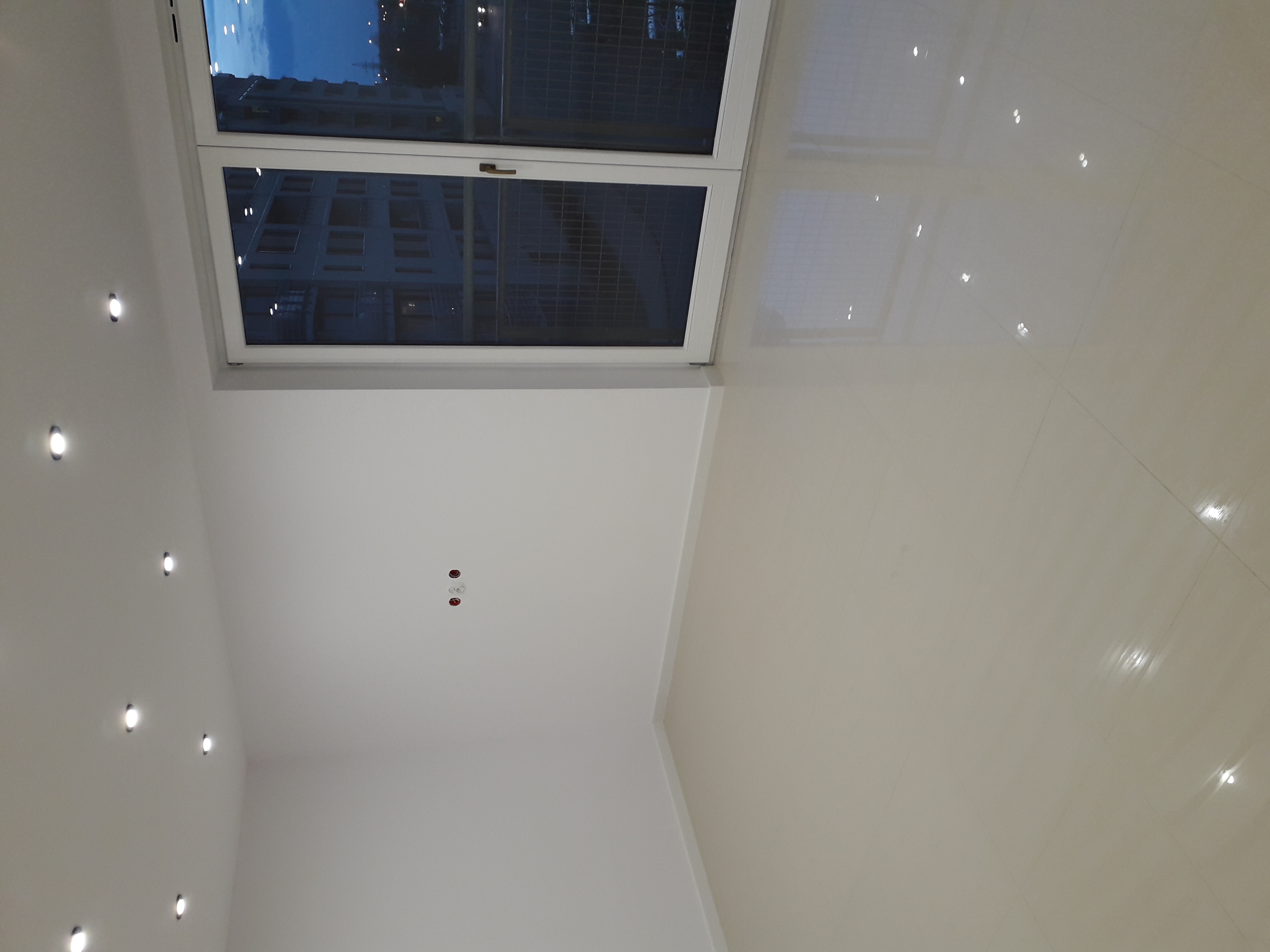 Widok z dołu na świeżo wykończone, jasne wnętrze z błyszczącą, beżową podłogą z płytek, białymi ścianami z zamontowanymi punktowymi lampami LED i oknem z widokiem na budynek po zmroku.