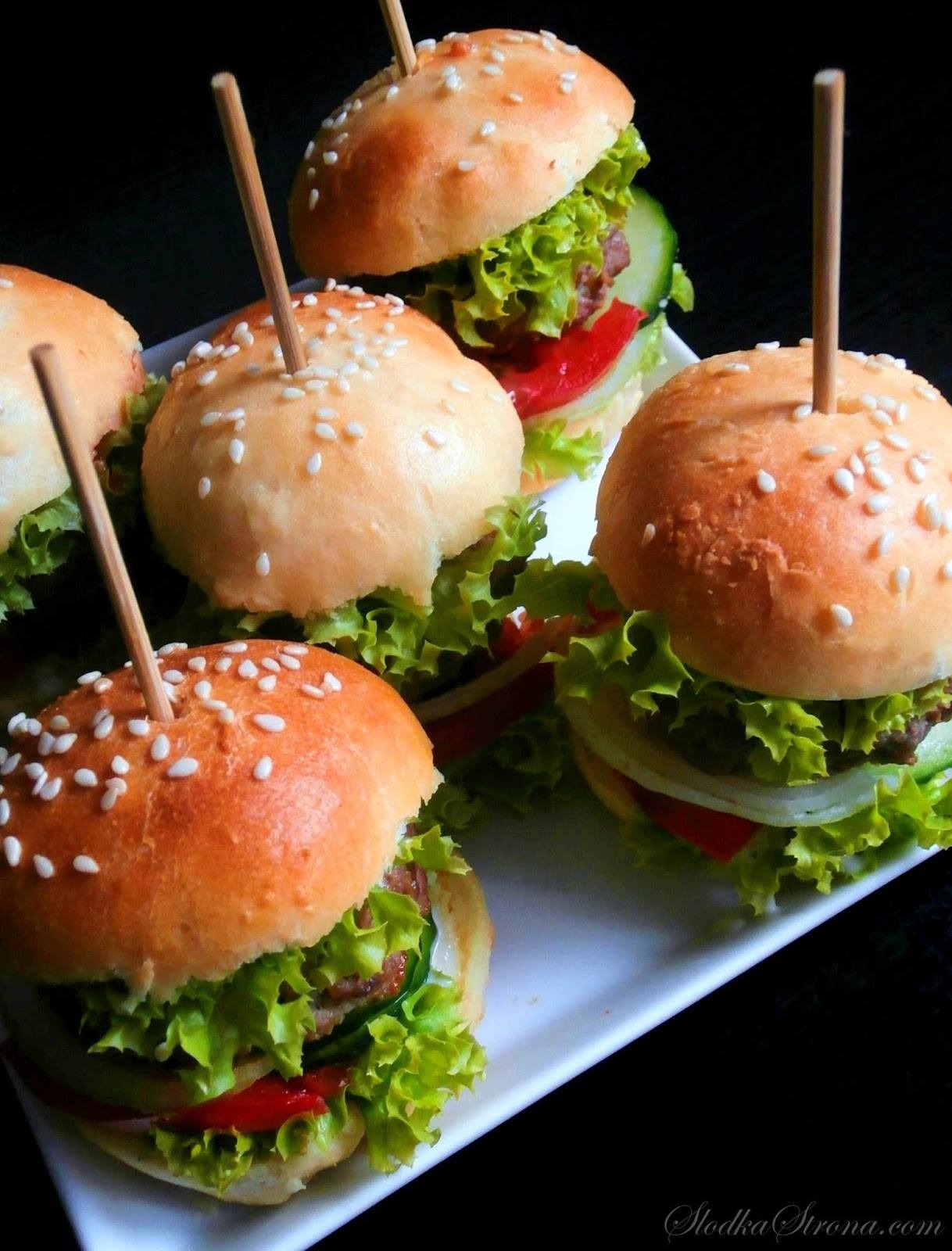 Miniaturowe burgery z sezamem, sałatą, pomidorem i ogórkiem na białej tacy, zabezpieczone wykałaczkami, z ciemnym tłem.