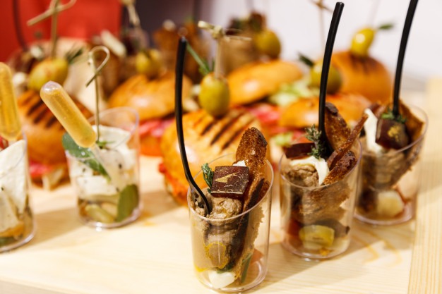 Elegancko podane przekąski w małych przezroczystych kubeczkach, z mini-burgerami w tle, na drewnianej tacy.