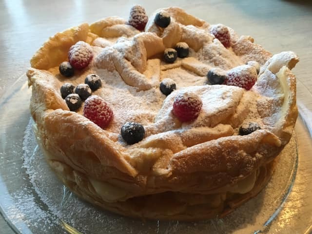 Tort z ciasta francuskiego przełożony kremem i udekorowany świeżymi malinami i borówkami, posypany cukrem pudrem, przygotowany w Gdyni.