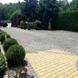 Ułożona z kostki brukowej posesja z geometrycznym wzorem, z obrzeżami z żółtej kostki, otoczona zadbaną zielenią i żywopłotem.