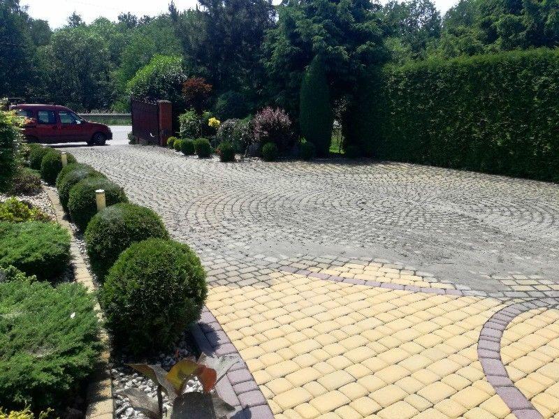 Ułożona z kostki brukowej posesja z geometrycznym wzorem, z obrzeżami z żółtej kostki, otoczona zadbaną zielenią i żywopłotem.