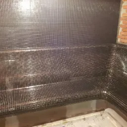 Wykończona sauna z ciemnymi, błyszczącymi mozaikami na ścianach i siedziskach, z fragmentem ściany wyłożonej czerwoną cegłą.