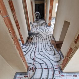 POL BAU STOKLOSA SPÓŁKA Z OGRANICZONĄ ODPOWIEDZIALNOŚCIĄ - Montaż ogrzewania podłogowego w korytarzu domu; widoczne rozłożone rury na izolacji z siatką, pracownik w oddali, surowe ściany z ościeżnicami.