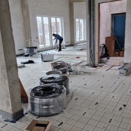 POL BAU STOKLOSA SPÓŁKA Z OGRANICZONĄ ODPOWIEDZIALNOŚCIĄ - Wnętrze w stanie surowym z widocznymi rolkami rur grzewczych na podłodze z izolacją, instalator w tle przy oknie, pionowe rury instalacyjne przy ścianie.