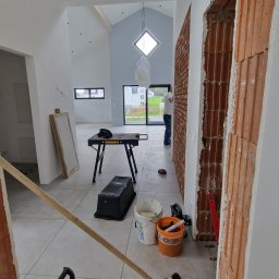 POL BAU STOKLOSA SPÓŁKA Z OGRANICZONĄ ODPOWIEDZIALNOŚCIĄ - Wnętrze domu w trakcie wykańczania, widoczne narzędzia glazurnicze, płytki podłogowe z krzyżykami dystansowymi, ceglana ściana i osoba w tle.