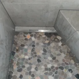 Wykończona podłoga kabiny prysznicowej z mozaiką z otoczaków w odcieniach szarości i beżu, obramowana płytkami ceramicznymi w kolorze szarym i odpływem liniowym.