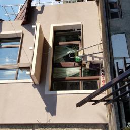 POL BAU STOKLOSA SPÓŁKA Z OGRANICZONĄ ODPOWIEDZIALNOŚCIĄ - Elewacja domu po malowaniu w odcieniach beżu, z brązowymi ramami okiennymi i widoczną drabiną oraz akcesoriami malarskimi na balkonie.