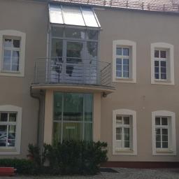 POL BAU STOKLOSA SPÓŁKA Z OGRANICZONĄ ODPOWIEDZIALNOŚCIĄ - Elewacja dwupiętrowego budynku z beżową fasadą, białymi obramowaniami okien, balkonem z przeszkloną werandą i widocznym fragmentem rusztowania oraz kontenera budowlanego z prawej strony.