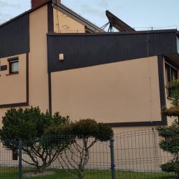 POL BAU STOKLOSA SPÓŁKA Z OGRANICZONĄ ODPOWIEDZIALNOŚCIĄ - Odnowiona elewacja domu z ciemnymi elementami wykończeniowymi i beżową fasadą. Widoczne okna, ogrodzenie i zadbana zieleń wokół budynku.