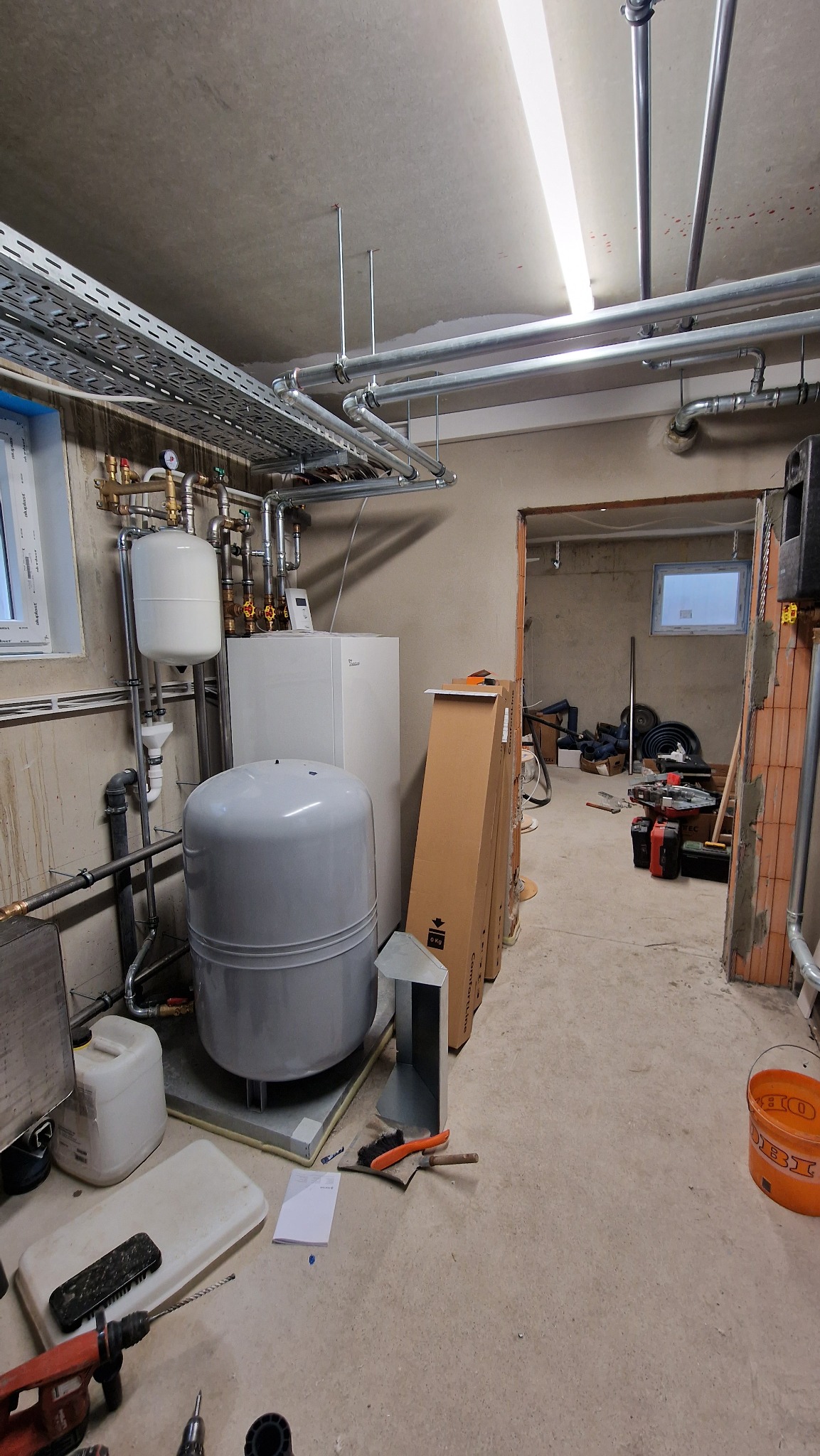 Piwnica w trakcie montażu instalacji hydraulicznej: zbiorniki, rury, narzędzia, kartony z nowymi urządzeniami w tle.