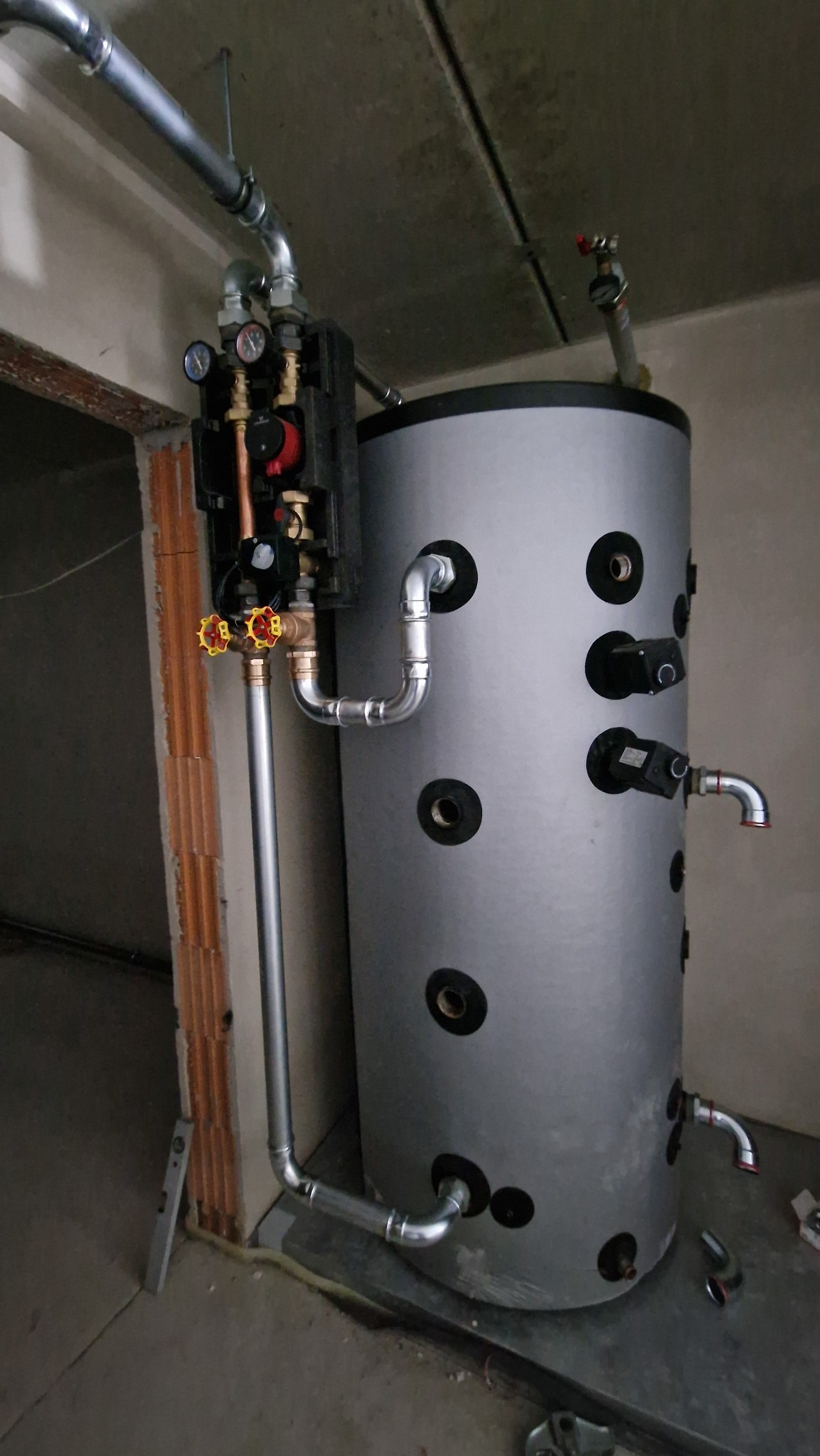 Szary bojler z licznymi przyłączami i zaworami, zamontowany w surowym pomieszczeniu z widoczną cegłą dziurawką i rurami instalacyjnymi.