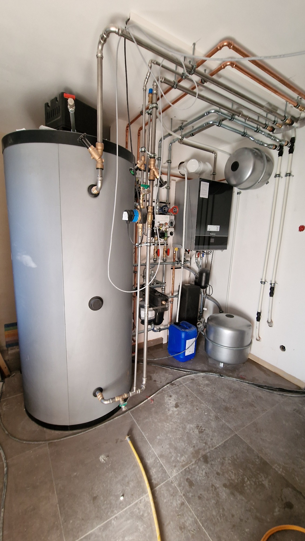Srebrny zasobnik ciepłej wody użytkowej z licznymi rurami miedzianymi i stalowymi, zaworami oraz osprzętem hydraulicznym na tle białej ściany i brązowej podłogi z płytek.