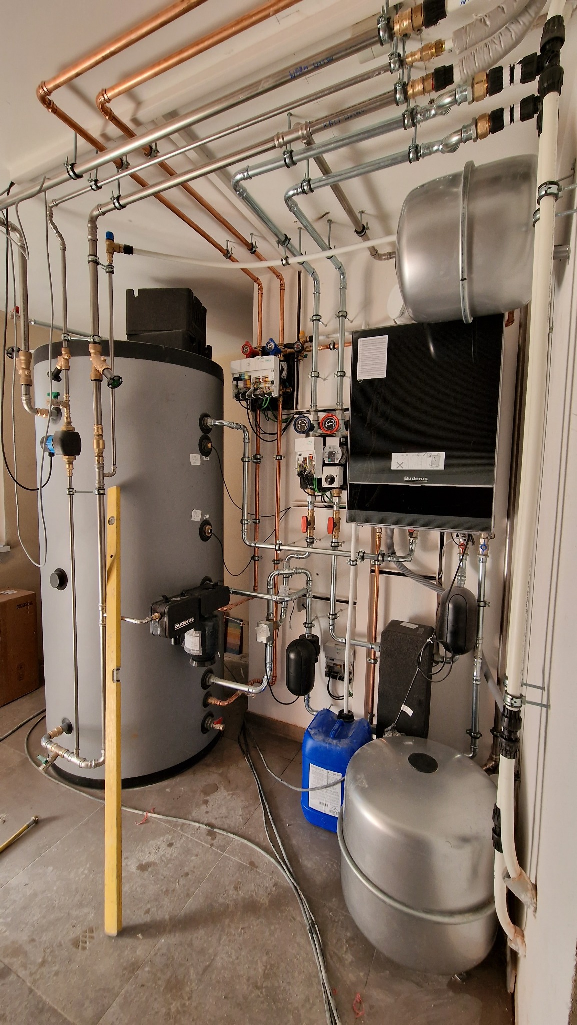 Złożona instalacja kotłowni z dwoma zbiornikami, kotłem Buderus, licznymi rurami miedzianymi i stalowymi, zaworami oraz osprzętem, widoczna poziomica i niebieski kanister.