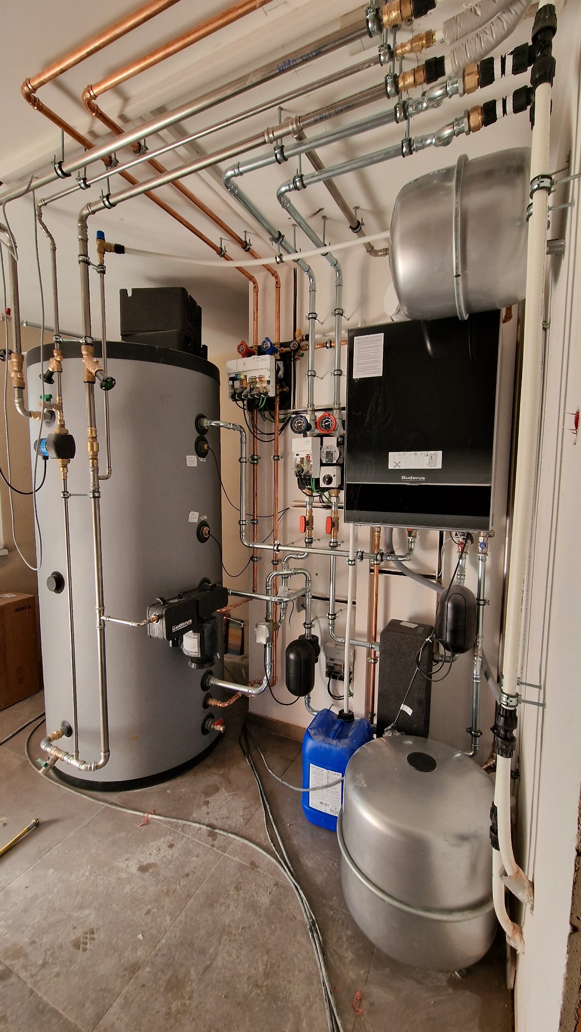 Kompleksowa instalacja kotła grzewczego Buderus z zasobnikiem wody, licznymi rurami miedzianymi i stalowymi oraz osprzętem, widok na kotłownię z niebieskim kanistrem i przewodami na podłodze.