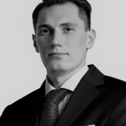 Portret młodego mężczyzny w czarnym garniturze i wzorzystym krawacie, na jasnym tle, z lekko zmrużonymi oczami.