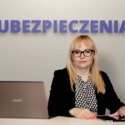 Kobieta w okularach siedzi za biurkiem przed laptopem i tabletem, w tle napis 'Ubezpieczenia' na ścianie.