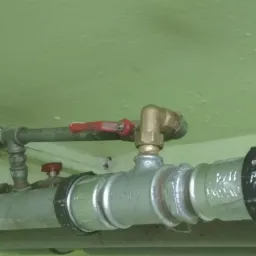 Szara rura z zaworem kulowym z czerwoną rączką i mosiężnym kolankiem, mocowana do sufitu pomalowanego na bladozielony kolor.