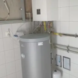 Szary zasobnik ciepłej wody użytkowej z białym piecem gazowym kondensacyjnym zamontowanym na ścianie wyłożonej białymi płytkami. Widoczne rury i zawory. Surowy stan ściany z cegły i instalacji...