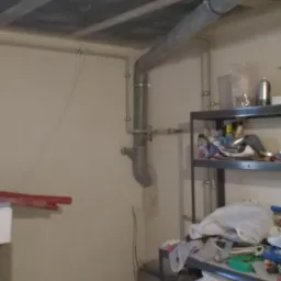 Szare rury kanalizacyjne zamontowane pod betonowym sufitem w piwnicy, widoczne obejmy mocujące i trójnik, na ścianie białe rurki instalacyjne, w tle regał z narzędziami i akcesoriami.