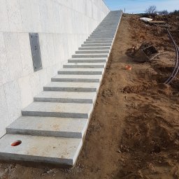 PHU TOMASZ TURCZYK - Granitowe schody zewnętrzne prowadzące na wzgórze, z widocznym przyłączem gazowym na ścianie i elementami robót ziemnych w tle. Solidne wykonanie, jasna kolorystyka.