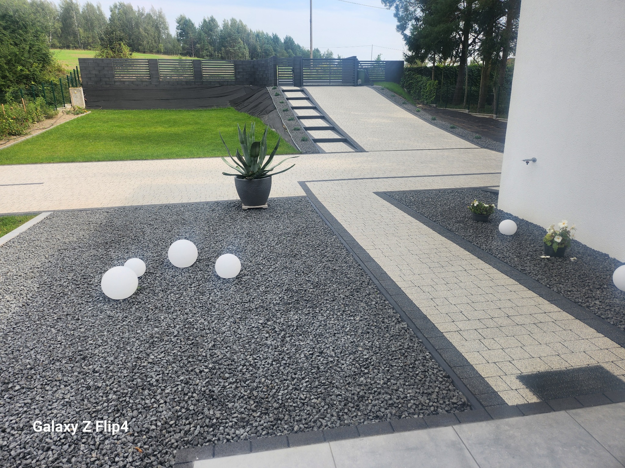 Nowoczesny podjazd z kostki brukowej w geometrycznym wzorze, otoczony szarym żwirem i dekoracyjnymi kulami. W tle widoczny trawnik i ogrodzenie.