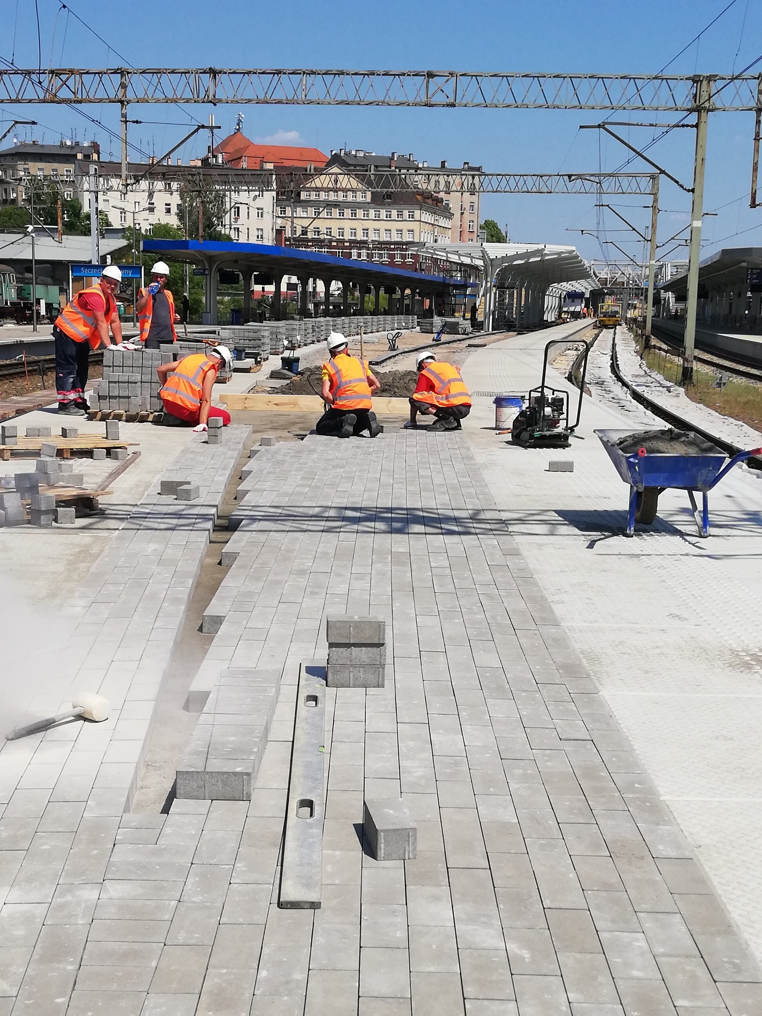 Układanie szarej kostki brukowej na peronie kolejowym. Pracownicy w pomarańczowych kamizelkach i białych kaskach. W tle budynki miejskie i infrastruktura kolejowa.