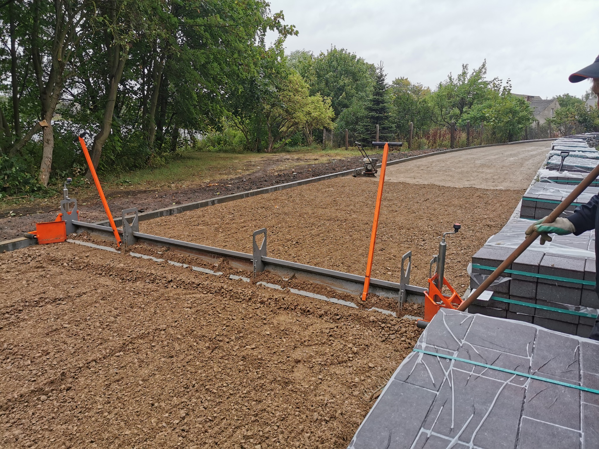 Przygotowanie podłoża pod kostkę brukową z użyciem narzędzi do poziomowania. W tle widoczny teren zielony i ułożona kostka brukowa.