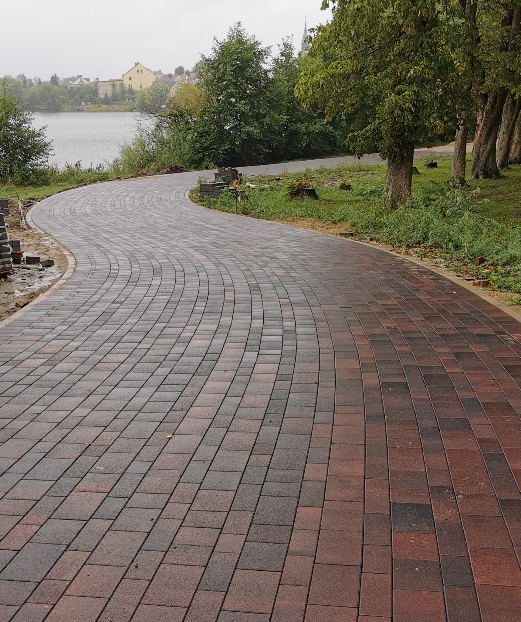Kręta ścieżka z kostki brukowej w odcieniach czerwieni i brązu, prowadząca wzdłuż jeziora w pochmurny dzień. Częściowo ułożona kostka, widoczne drzewa i budynki w tle.