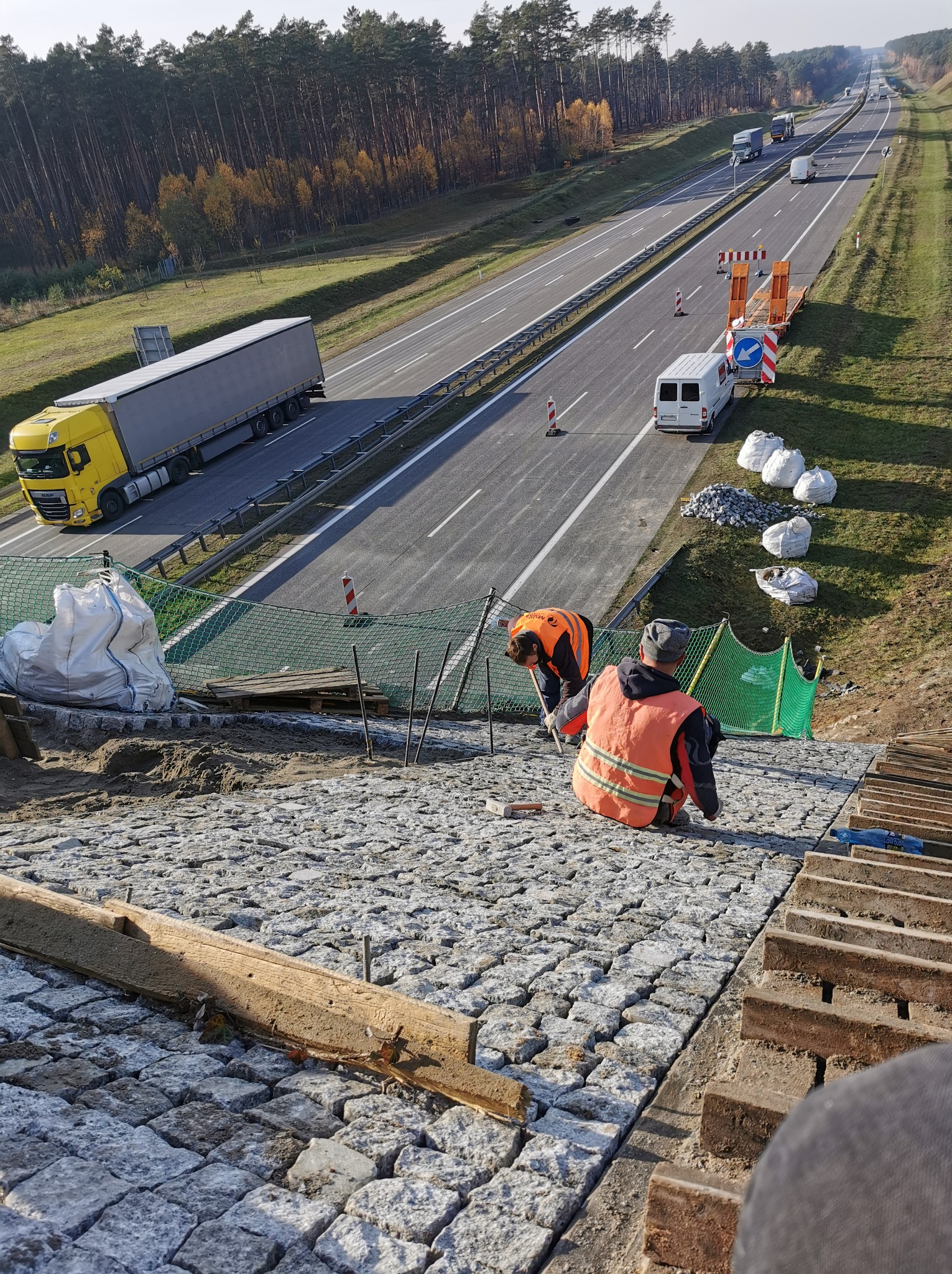 Drogowcy układają kostkę brukową na poboczu autostrady. Widoczne narzędzia, worki z materiałem i ruch samochodowy w tle. Praca w toku, dzień.