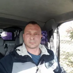 Mężczyzna w samochodzie, ubrany w kurtkę z szarymi wstawkami i niebieską koszulkę. Widoczne siedzenia, pas bezpieczeństwa i fragment krajobrazu za oknem.