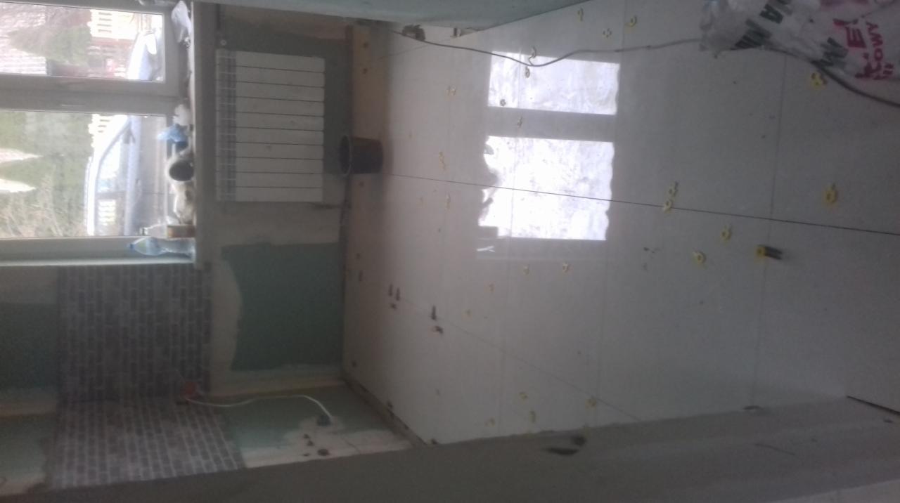 Układanie błyszczących, jasnych płytek podłogowych z użyciem krzyżyków dystansowych, widoczna instalacja grzejnika i fragment okładziny ściennej z cegiełek.