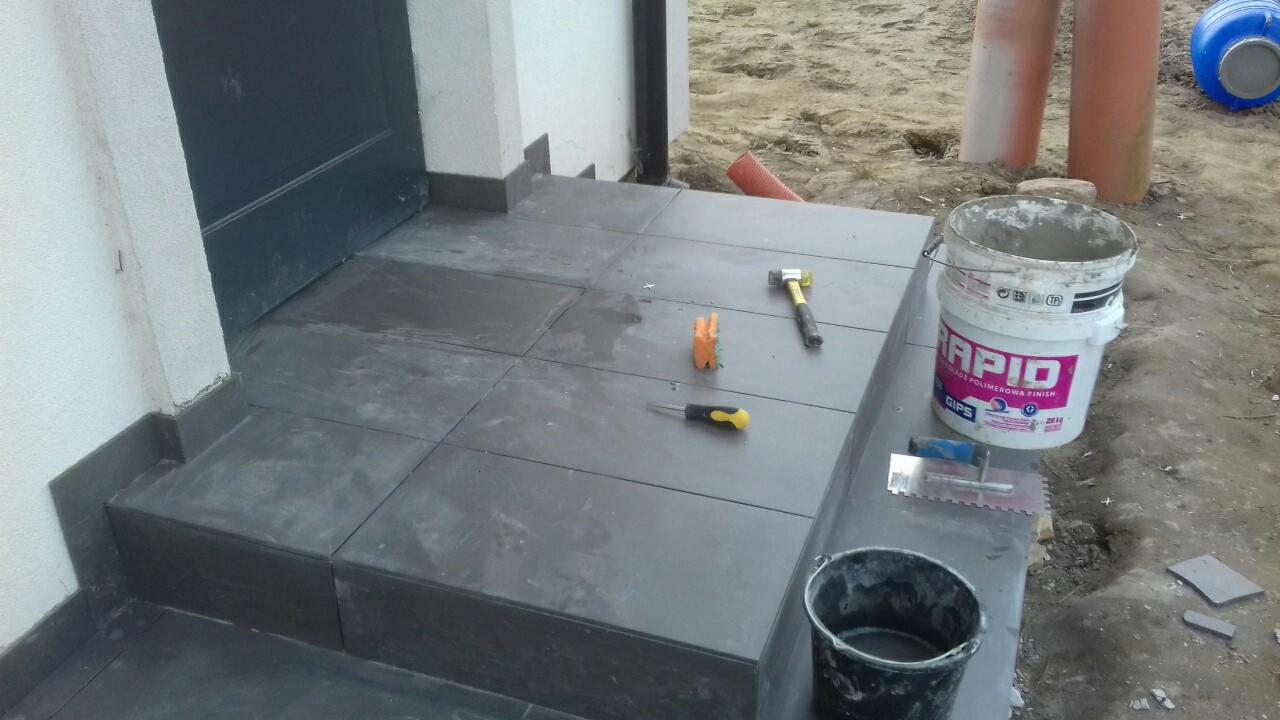 Wyłożone szarymi płytkami schody wejściowe do budynku, widoczne narzędzia: młotek, śrubokręt, wiadro z zaprawą Rapid, kielnia ząbkowana, czarne wiadro, fragment rynny i rury kanalizacyjne w tle.