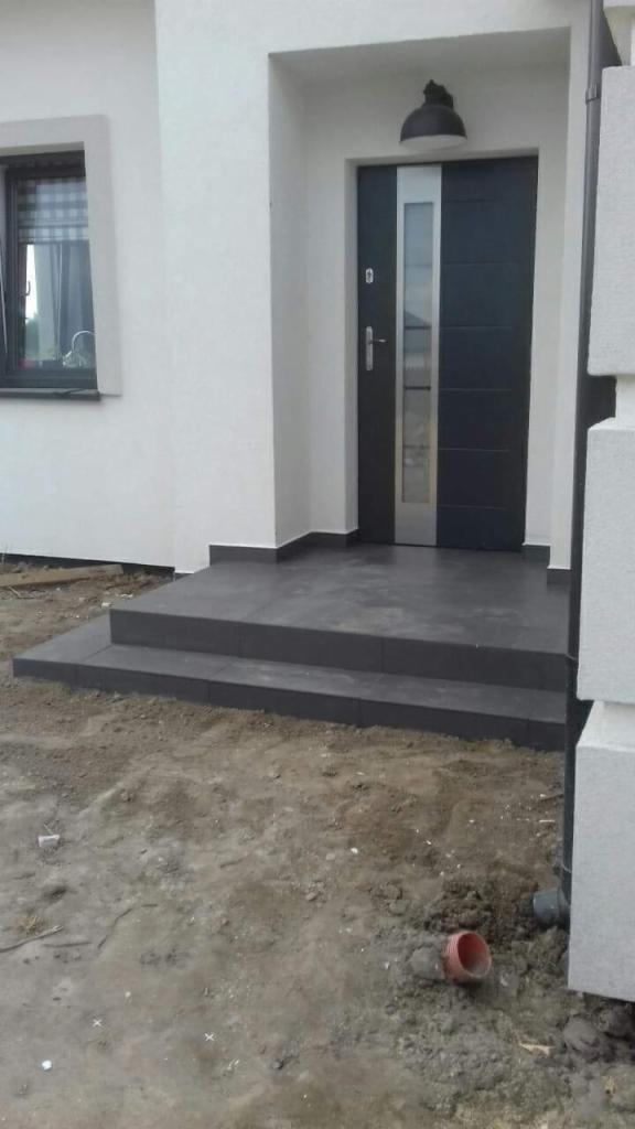 Nowoczesne wejście do domu z ciemnoszarymi płytkami na schodach i podestem, czarne drzwi z pionowym przeszkleniem, lampa zewnętrzna nad drzwiami, teren wokół jeszcze niezagospodarowany, widoczna...