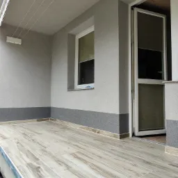 Wykończony balkon z podłogą imitującą drewno, szare ściany z ciemniejszym cokołem i otwarte białe drzwi balkonowe.