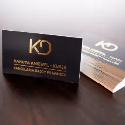 Eleganckie wizytówki Kancelarii Radcy Prawnego Danuty Kniewel-Kurek, z logo KD w złotym kolorze na ciemnym tle, ułożone na drewnianym biurku.