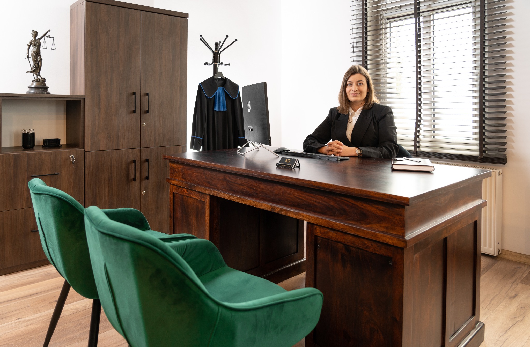 Kobieta w biurze prawnym siedzi za biurkiem z togą adwokacką w tle. Na biurku wizytówka, notatnik i komputer. W tle szafa i figura Temidy.