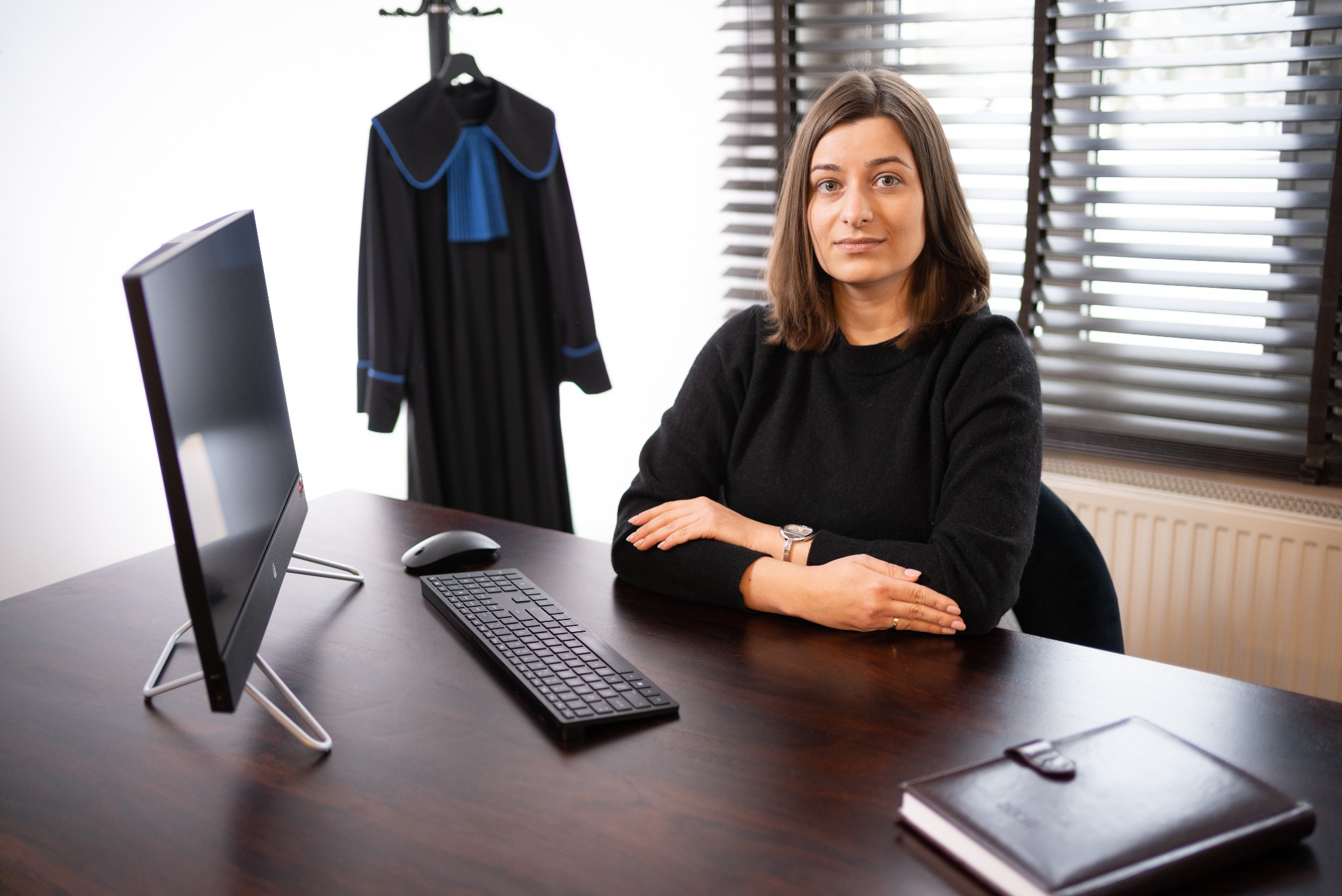 Portret kobiety adwokat przy biurku z komputerem, w tle toga na wieszaku. Ujęcie w biurze, z naturalnym światłem wpadającym przez żaluzje.