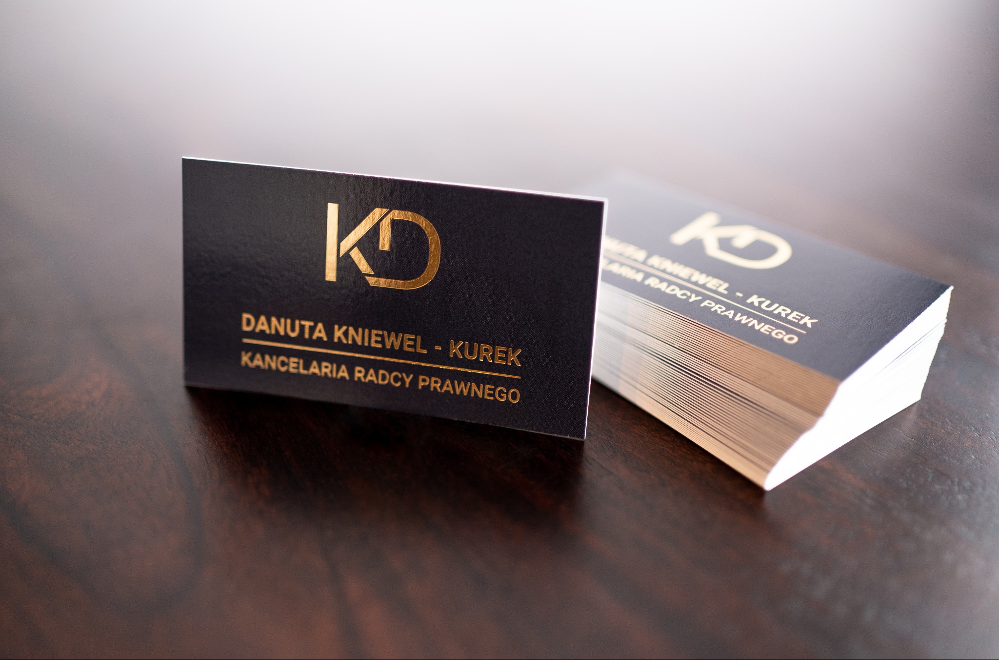Eleganckie wizytówki Kancelarii Radcy Prawnego Danuty Kniewel-Kurek, z logo KD w złotym kolorze na ciemnym tle, ułożone na drewnianym biurku.